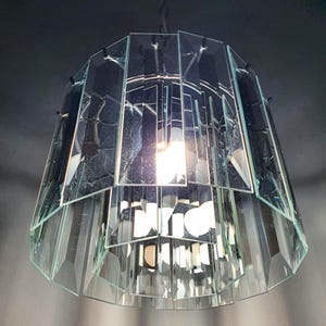 Puede incluir: Una lámpara colgante cilíndrica construida con múltiples paneles de vidrio verticales y rectangulares dispuestos en dos niveles superpuestos. Las piezas de vidrio transparente están montadas sobre un marco de metal oscuro, con una bombilla visible.