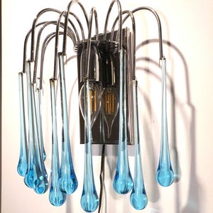 May include: A modern wall sconce with a chrome finish and blue glass teardrop-shaped pendants. The sconce has two light bulbs.