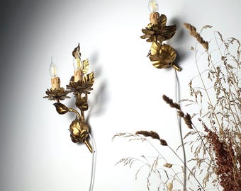 May include: Two gold-colored metal wall sconces featuring floral and leaf motifs. One sconce has two light sockets with candle-shaped bulbs, while the other has one. Both fixtures include white electrical cords extending from the base.