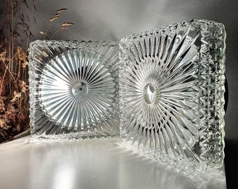 May include: Two square glass light fixtures featuring a clear, pressed pattern with a central circular indentation and radiating sunburst lines. The edges of the glass panels have a textured, geometric grid design that catches the light.