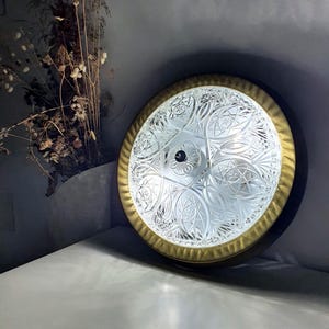 Könnte beinhalten: Eine runde Deckenleuchte mit einem klaren, gepressten Glasschirm, der ein geometrisches Stern- und Blumenmuster aufweist. Das Glas ist von einem breiten, strukturierten messingfarbenen Metallrand mit dunkler Außenkante und einem schwarzen Knopf eingefasst.