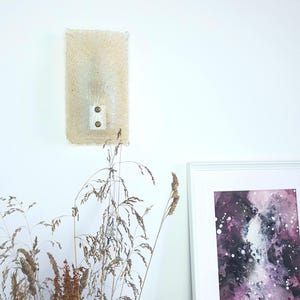 Op de afbeelding: Een witte wandlamp met een getextureerde glazen kap en twee messing schroeven. De lamp is gemonteerd op een witte muur en wordt verlicht door een gloeilamp. Op de voorgrond staat een boeket gedroogde grassen in een vaas.