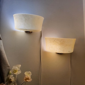 May include: Two wall-mounted sconces with a soft, textured appearance. The sconces have a bowl-like shape and emit a warm, diffused light. The fixtures are mounted on a neutral-colored wall, with visible power cords. The overall aesthetic is minimalist and elegant.