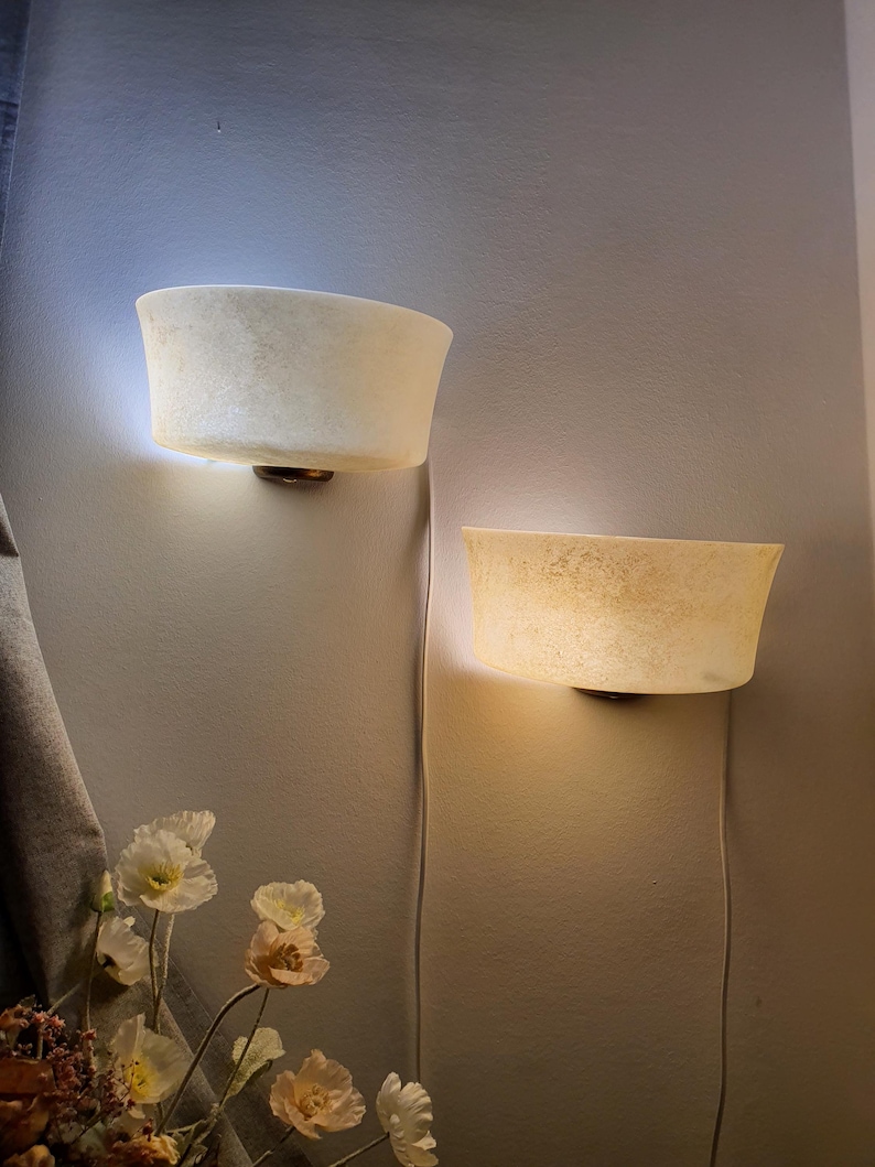 May include: Two wall-mounted sconces with a soft, textured appearance. The sconces have a bowl-like shape and emit a warm, diffused light. The fixtures are mounted on a neutral-colored wall, with visible power cords. The overall aesthetic is minimalist and elegant.