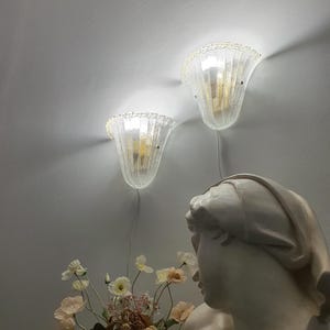 Könnte beinhalten: Zwei Wandleuchten mit klaren, strukturierten Glasschirmen in geriffelter Glockenform. Die Lampen strahlen ein warmes Licht an die Wand ab. Darunter befinden sich eine weiße Büste eines Kopfes und ein Strauß aus blassrosa und weißen Kunstblumen.