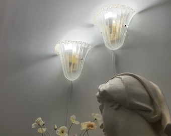 May include: Two wall-mounted sconces featuring clear, textured glass shades with a fluted, bell-like shape. The lights emit a warm glow against the wall. Below, a white sculpted bust of a head and a bouquet of pale pink and white artificial flowers.