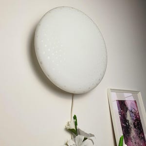 May include: A white, round, wall-mounted light fixture with a dotted pattern. The fixture is connected to a white cord. A framed picture and white flowers are visible below the light.