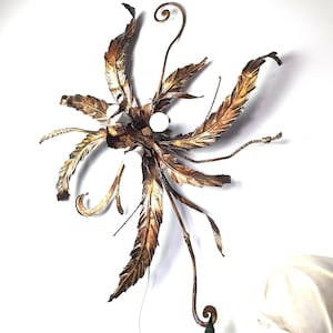 May include: A wall-mounted light fixture crafted from metal with a bronze and gold finish, featuring multiple elongated, textured leaf-shaped elements arranged in a radial pattern. The design includes three small, round, white light bulbs.