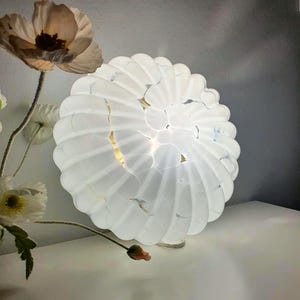 May include: A white, spherical lamp with a textured, petal-like design. The lamp is illuminated from within, casting a soft glow. Dried flowers in neutral tones are arranged nearby, suggesting a home decor theme.