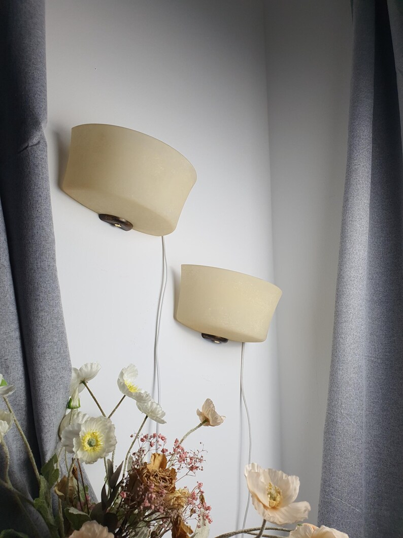 May include: Two wall-mounted sconces featuring semi-circular, frosted cream-colored glass shades. Each shade is attached to a dark brown circular wooden base. A thin white electrical cord hangs vertically from the bottom of each light fixture.