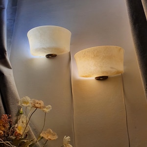 May include: Two wall sconces with a textured, off-white shade and a dark brown base are mounted on a light-colored wall. The sconces are illuminated, casting a warm glow. The scene is framed by a curtain and floral arrangement.