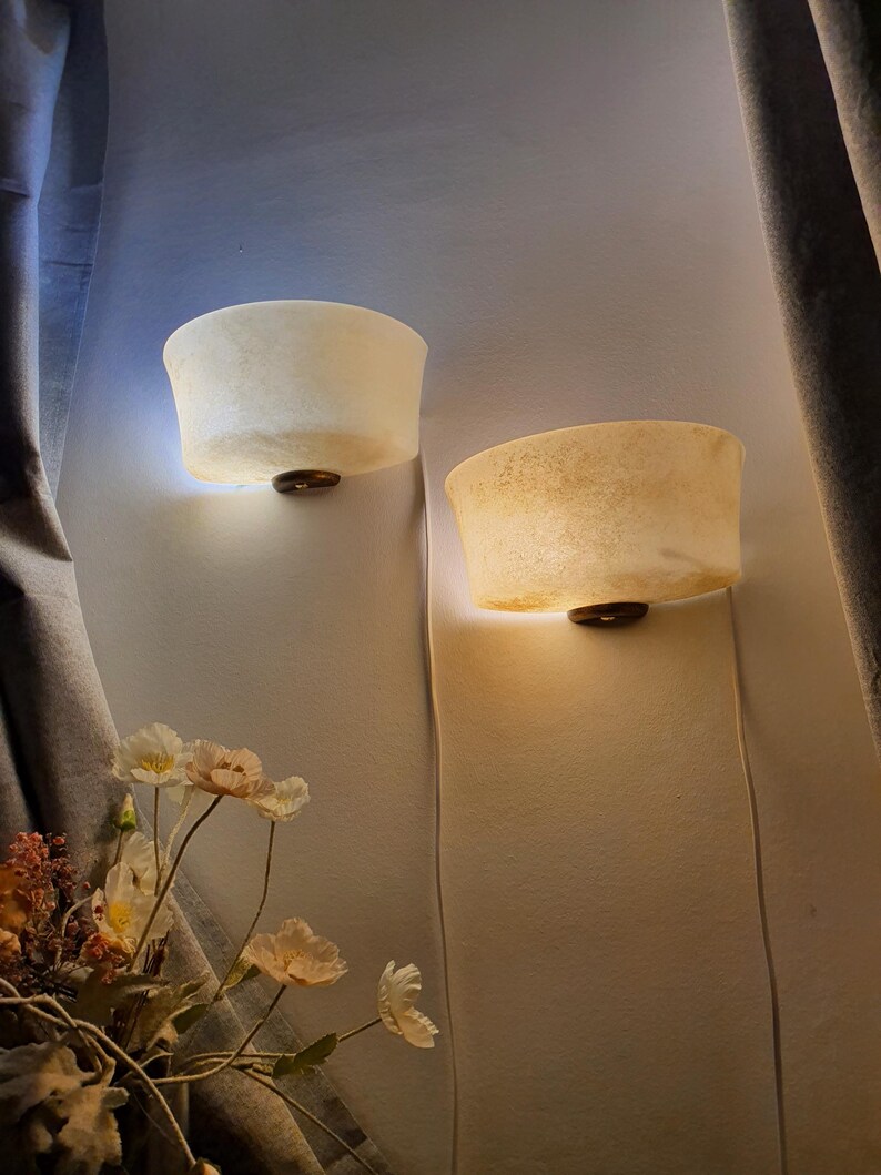 May include: Two wall sconces with a textured, off-white shade and a dark brown base are mounted on a light-colored wall. The sconces are illuminated, casting a warm glow. The scene is framed by a curtain and floral arrangement.
