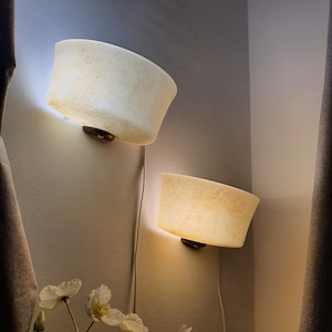 May include: Two wall-mounted sconces featuring semi-circular, off-white, textured shades that emit a warm, diffused glow. Each lamp is attached to the wall with a dark circular base and includes a white power cord hanging downward.