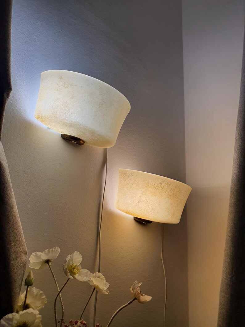 May include: Two wall-mounted sconces featuring semi-circular, off-white, textured shades that emit a warm, diffused glow. Each lamp is attached to the wall with a dark circular base and includes a white power cord hanging downward.
