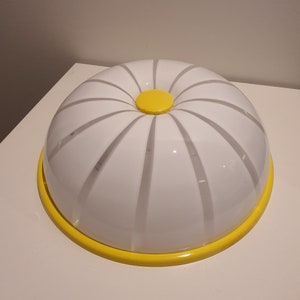 May include: A white plastic food cover with a yellow rim and a yellow center knob. The cover has a ribbed design.
