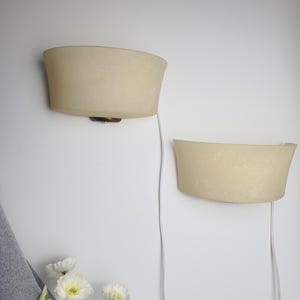 May include: Two semi-circular wall sconces with frosted, off-white glass shades. Each lamp features a dark metal base and a white electrical cord extending downward. The fixtures are mounted on a plain wall, positioned at different heights.