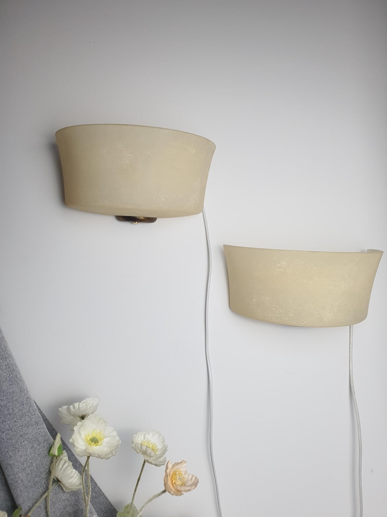 May include: Two semi-circular wall sconces with frosted, off-white glass shades. Each lamp features a dark metal base and a white electrical cord extending downward. The fixtures are mounted on a plain wall, positioned at different heights.