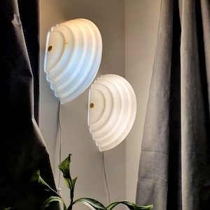 Peut inclure: Deux lampes murales en forme de coquillage, dégageant une lumière chaude et diffuse. Les lampes blanches présentent un design strié et un petit accent circulaire. Elles sont montées sur un mur de couleur claire, avec des rideaux sombres et de la verdure au premier plan.