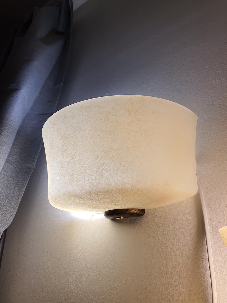 May include: A wall-mounted light fixture with a frosted, off-white shade. The shade has a curved shape and is mounted on a dark, circular base. The fixture is installed on a light gray wall.