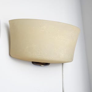 May include: A wall-mounted light fixture with a curved, off-white shade. The shade has a textured surface and is attached to a dark brown base. A white cord extends from the bottom of the fixture.