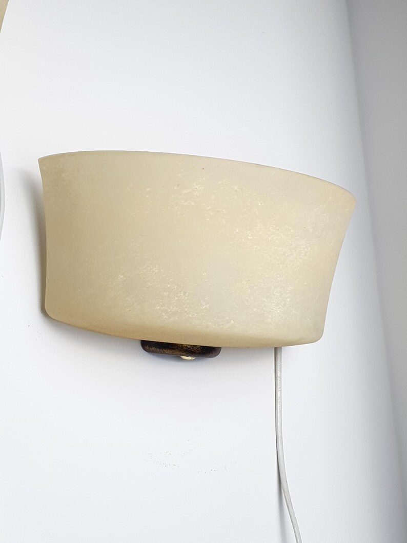 May include: A wall-mounted light fixture with a curved, off-white shade. The shade has a textured surface and is attached to a dark brown base. A white cord extends from the bottom of the fixture.