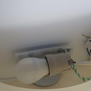 May include: A white LED light bulb installed in a gray ceramic socket, mounted on a metal plate. The electrical wiring includes a green and white striped cord. The light fixture is attached to a white surface.
