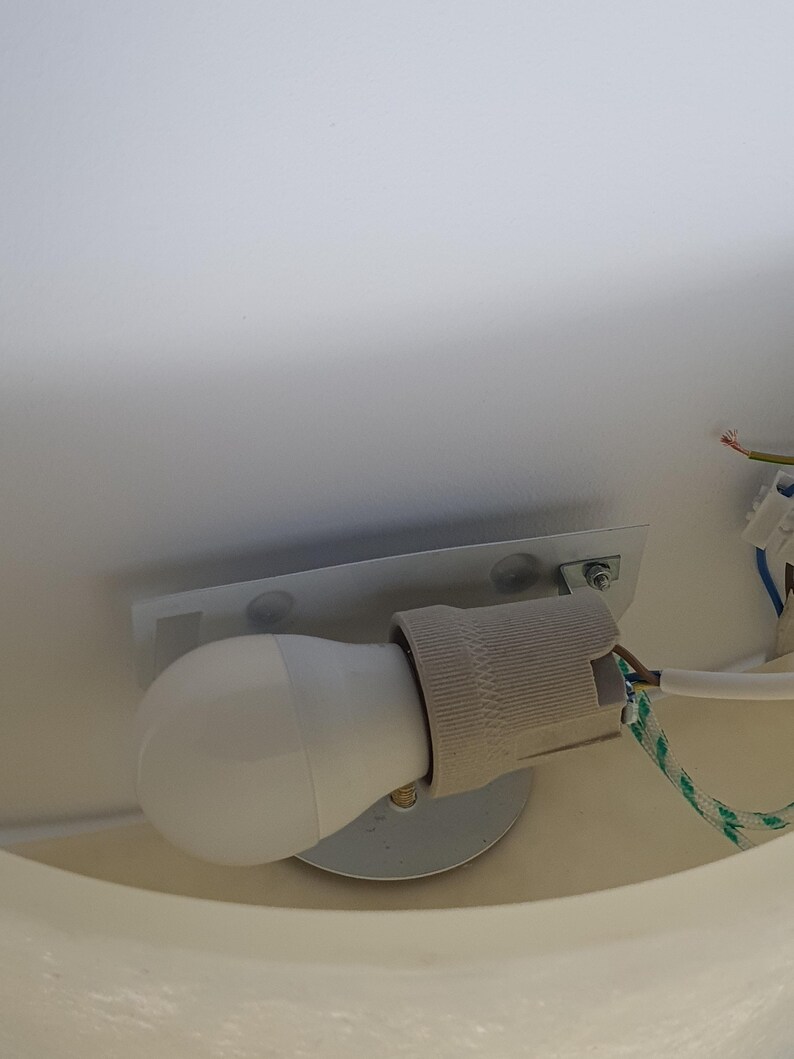May include: A white LED light bulb installed in a gray ceramic socket, mounted on a metal plate. The electrical wiring includes a green and white striped cord. The light fixture is attached to a white surface.