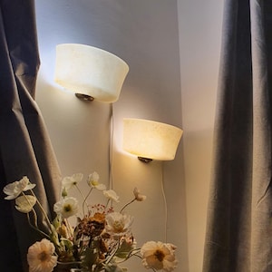 May include: Two wall-mounted sconces with a warm, diffused glow. The sconces have a curved, off-white shade and are mounted on a light-colored wall. A bouquet of flowers in a vase sits below the sconces, with a dark curtain on the side.