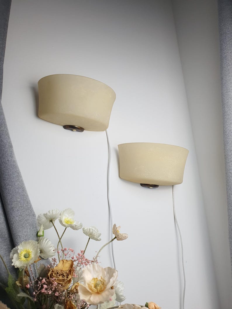 May include: Two cream-colored wall sconces with a curved design are mounted on a white wall. Each sconce has a dark brown base and a visible electrical cord. A bouquet of flowers is in the foreground.
