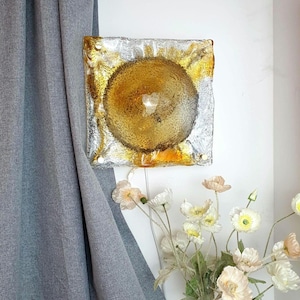 以下が含まれることがあります： 金と琥珀色の円形テクスチャデザインが施された正方形のウォールアート。 アートワークは白い壁に取り付けられ、灰色のカーテンと花瓶の隣にあります。 この作品は、ユニークで抽象的な外観をしています。