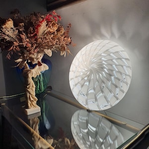 Könnte beinhalten: Eine kreisförmige Tischleuchte aus Glas mit einem weißen und transparenten Wirbelmuster, das vom Zentrum ausstrahlt. Die Lampe strahlt ein sanftes Licht aus, erzeugt einen deutlichen Spiraleffekt auf ihrer Oberfläche und steht aufrecht auf einer Glasfläche.