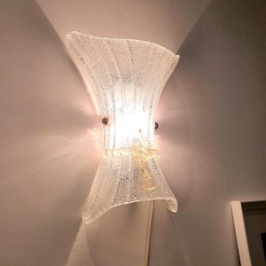 May include: A clear glass wall sconce with a textured surface and a gold metal base. The sconce is lit with a warm white light bulb.