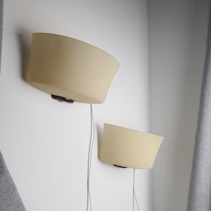 May include: Two cream-colored wall sconces with a curved design are mounted on a white wall. Each sconce has a dark brown base and a thin, silver-colored wire. The sconces are positioned at different heights.