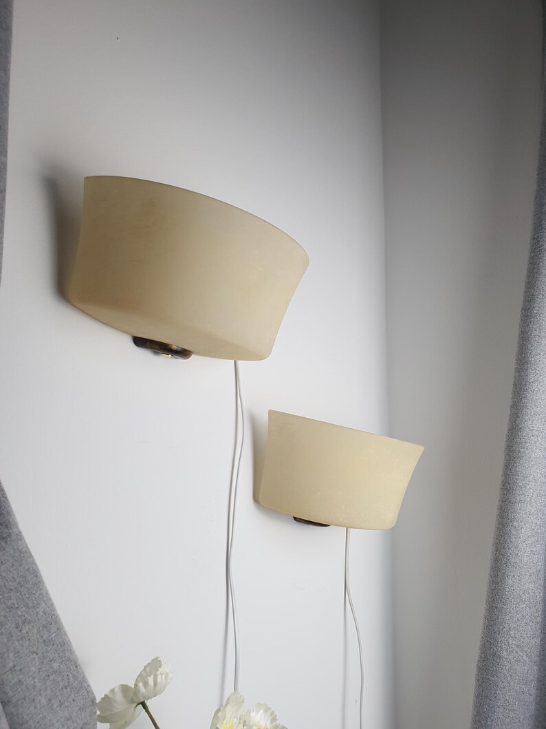 May include: Two cream-colored wall sconces with a curved design are mounted on a white wall. Each sconce has a dark brown base and a thin, silver-colored wire. The sconces are positioned at different heights.