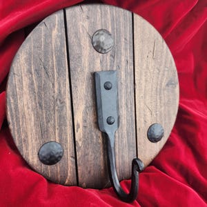 May include: A round, dark stained wooden wall hook with a black metal hook and decorative rivets. The wooden base is made of vertical planks and has a rustic, handcrafted appearance. The hook is mounted on a red background.