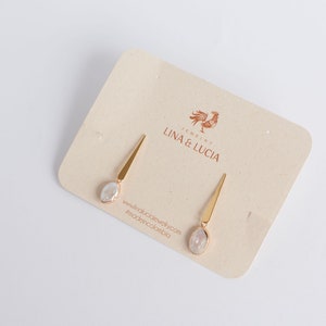 Puede incluir: Un par de pendientes de oro con una piedra blanca nacarada en cada pendiente. Los pendientes se muestran en una tarjeta con el texto "Jewelry Lina & Lucia" y un logotipo de gallo.