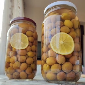 Può includere: Due barattoli di vetro pieni di olive verdi e salamoia. I barattoli hanno coperchi bianchi e sono appoggiati su una superficie marrone chiaro. È visibile una fetta di limone in ogni barattolo.