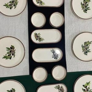 Può includere: Una collezione di piatti e vassoi in ceramica bianca con bordo marrone e design a ramo d'ulivo. Il set comprende piatti rotondi, vassoi ovali e piccole ciotole rotonde. Il design del ramo d'ulivo presenta foglie verdi e olive nere.
