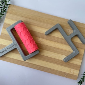 May include: A gray roller with a pink honeycomb pattern and a gray stand are displayed on a bamboo cutting board. The roller has the text "PRINT AND CUT CO." printed on the handle. Green leafy sprigs are in the background.