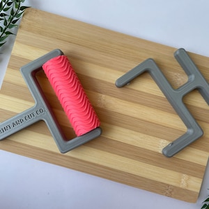 May include: A gray print roller with a pink, wavy patterned roller and a gray handle is on a wooden cutting board. The text "PRINT AND CUT CO." is printed on the handle. A gray tool is also on the board. Green leafy sprigs are in the background.