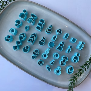 Puede incluir: Una colecci&oacute;n de cortadores de galletas de pl&aacute;stico azul en varias formas geom&eacute;tricas, incluyendo c&iacute;rculos, cuadrados, tri&aacute;ngulos y arcos, dispuestos en un plato de cer&aacute;mica gris claro.