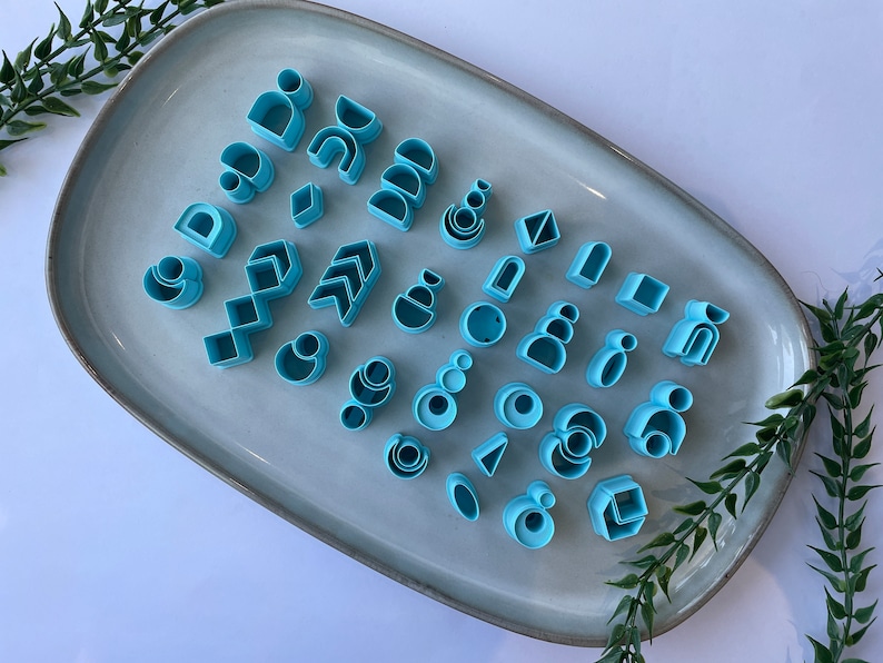 Puede incluir: Una colecci&oacute;n de cortadores de galletas de pl&aacute;stico azul en varias formas geom&eacute;tricas, incluyendo c&iacute;rculos, cuadrados, tri&aacute;ngulos y arcos, dispuestos en un plato de cer&aacute;mica gris claro.