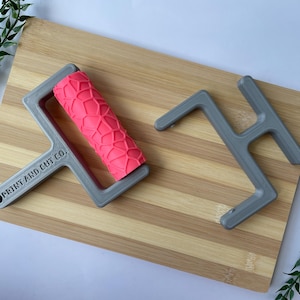 May include: A gray roller with a pink textured surface and a gray handle, next to a gray H-shaped tool. The roller has the text "PRINT AND CUT CO." on the handle. The items are on a wooden cutting board with green leafy sprigs.