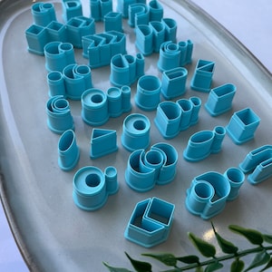 Puede incluir: Un juego de 30 cortadores de galletas de pl&aacute;stico azul en varias formas, incluyendo c&iacute;rculos, cuadrados, rect&aacute;ngulos, tri&aacute;ngulos y &oacute;valos. Los cortadores est&aacute;n dispuestos en un plato de cer&aacute;mica gris claro.