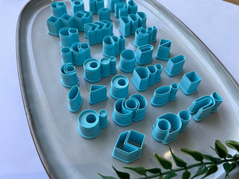 Puede incluir: Un juego de 30 cortadores de galletas de pl&aacute;stico azul en varias formas, incluyendo c&iacute;rculos, cuadrados, rect&aacute;ngulos, tri&aacute;ngulos y &oacute;valos. Los cortadores est&aacute;n dispuestos en un plato de cer&aacute;mica gris claro.