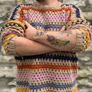 May include: A multi-coloured crocheted sweater with stripes of orange, pink, blue, and yellow. The sweater has a loose fit and a round neckline.