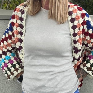 Könnte beinhalten: Ein handgefertigter Häkelcardigan mit Granny-Square-Design. Die Ärmel zeigen Quadrate in Rot-, Blau-, Lila- und Cremetönen. Der offene Cardigan scheint aus weichem Garn gefertigt zu sein.