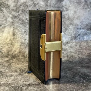 May include: A small, antique black book with gold-gilded edges and a gold clasp. The book has a textured cover and a blue ribbon bookmark. The edges of the pages are a warm, copper color.