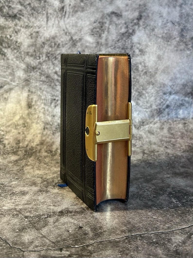 May include: A small, antique black book with gold-gilded edges and a gold clasp. The book has a textured cover and a blue ribbon bookmark. The edges of the pages are a warm, copper color.