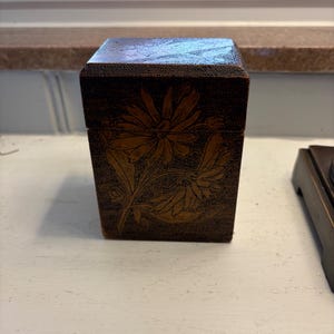 May include: A dark brown, rectangular box with a floral design. The box has a hinged lid and is decorated with a carved daisy and leaf pattern. The box is sitting on a white surface.